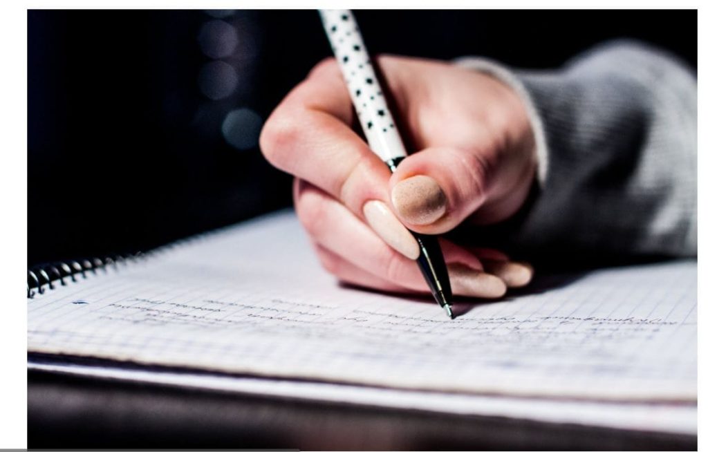 On voit une main d'élève écrivant une fiche de lecture sur une feuille de cahier avec un stylo.